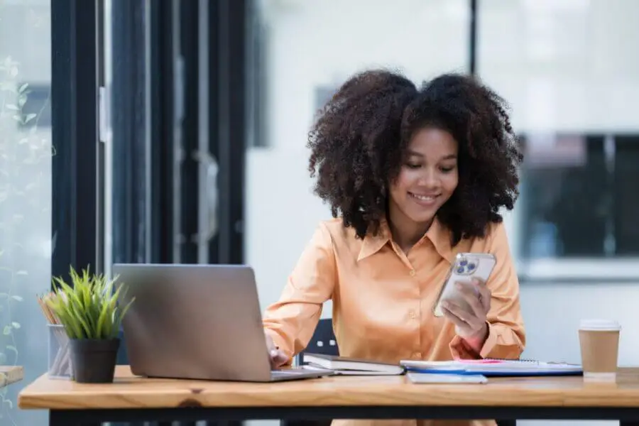 Portrait of a beautiful confident businesswoman using a laptop computer holding a mobile phone sitting in a modern office. Smiling African American freelancer working online from home.