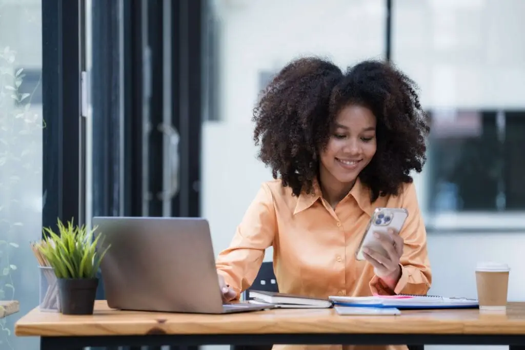 Young woman at her home office desktop, looking at her cell phone and laptop, smiling at her bank balance